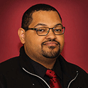 Kyle is pictured from the shoulders up. He has black hair, dark skin and is wearing glasses.