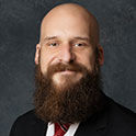 Richard is pictured from the neck up. He has pale skin, a dark brown beard, and no hair on his head. He is smiling. 