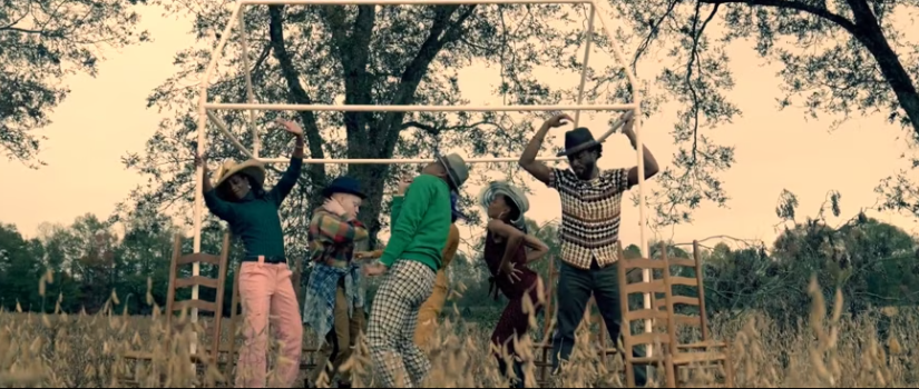 Six dancers perform in a field beneath a skeletal wooden frame resembling a small dance hall, surrounded by tall grass and trees, evoking a reconstructed historical space.