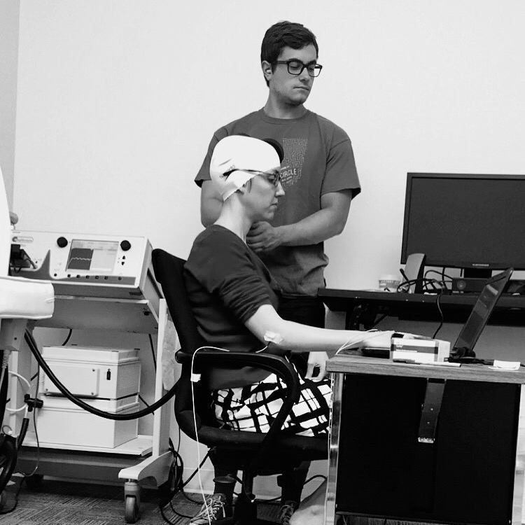 Person seated at a computer wearing a cap with sensors attached, connected to monitoring equipment, while another person stands behind them adjusting the setup in a lab environment.