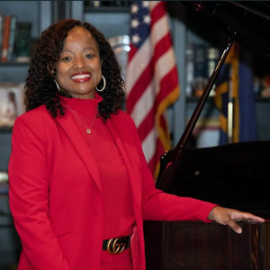DeAndrea Benjamin stands next to a piano with a bookcase and a U.S. flag in the background